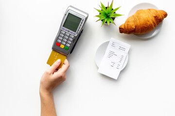 Hand is paying the bill by pos terminal in cafe or restaurant. Cup of black coffee and croissant, payment wireless machine and bill on white background, top view