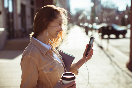 Smiling young woman using smartphone with earphones on city sidewalk
