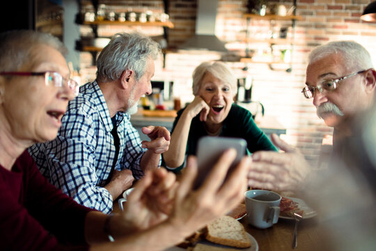 Happy senior friends laughing over smartphone at kitchen table