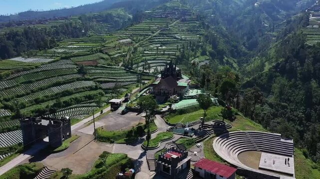 Aerial view of lush green tea plantations and a resort with an amphitheater nestled in a mountainous landscape.