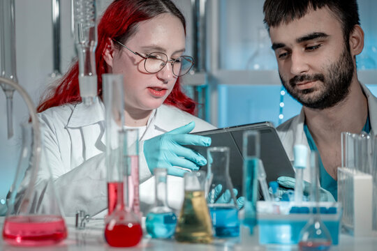 Young female lab technician working on a tablet while collaborating with male colleague in modern laboratory. Digital technology in chemistry research, teamwork, analysis and innovation concept. - Powered by Adobe