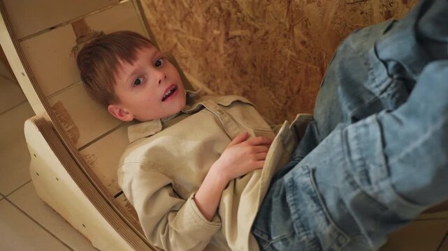 boy in cardboard pod reclining, cozy sweater and jeans, plywood backdrop, gentle smile frames show child exploring imagination, hiding in nook, resting like reader and daydreamer, soft indoor