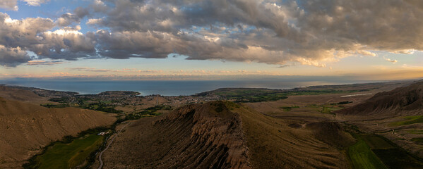 Expansive view of serene landscape featuring mountains, a large Issyk-Kul lake, and fields under dramatic clouds. Golden hour lighting enhances the peaceful atmosphere.