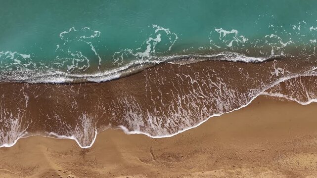 Golden sandy beach with white foamy waves crashing onto it. A bird's-eye view video of the sea with the soothing rhythm of the waves and its turquoise color.