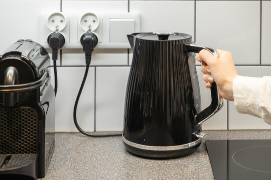 Person's hand grasping stylish black electric kettle on a modern kitchen counter, beside a coffee machine, highlighting morning routine and household energy use