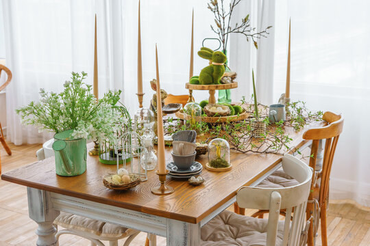 Stylish Easter table decor with candles, greenery, bunny figurines and natural elements in bright spring room interior. Cozy festive dining setup with wooden table, soft daylight and seasonal details