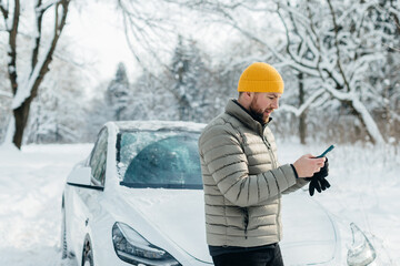 Driver checking smartphone near electric car in winter
