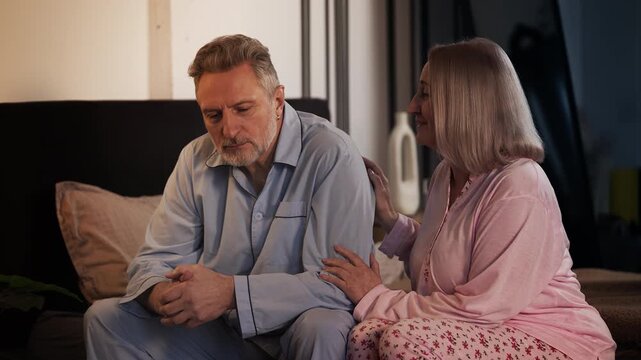 Loving middle-aged wife supporting upset husband while sitting on bed, trust