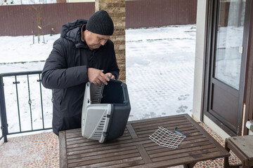Adult man in warm winter clothing adjusting plastic pet carrier on outdoor terrace with snow around.