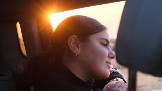 Happy beautiful teenage girl looks out of a car window with surprised and admiring eyes at beautiful nature and a sunny sunset while driving through wonderful natural places. Opening new horizons
