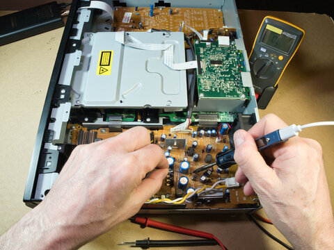 Close-up of technician's hands using a multimeter to diagnose or repair complex electronic circuitry inside an open DVD/Blu-ray player or similar device.