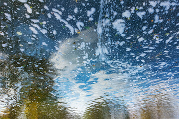 Fototapeta premium A lake background with patches of foam reflecting a blue sky, with clouds and autumn trees distorted by gentle ripples.