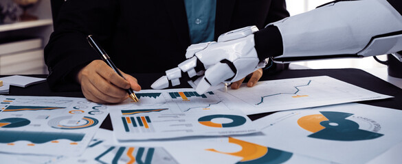 A human and a robotic hand engage in data analysis during a business meeting, showcasing the integration of technology in modern workplaces through charts and graphs.Noogenesis
