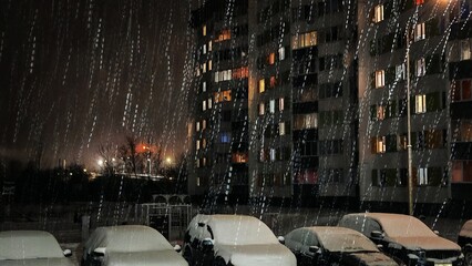 Heavy Night Snowfall Over Urban Residential Area. Dramatic winter night scene with heavy snowfall falling over snow-covered parked cars and multi-story apartment buildings.