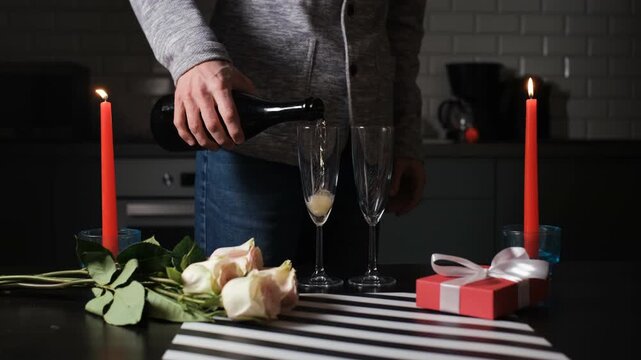 A man prepares for Valentines Day by pouring champagne into glasses while candles light a table. Roses and a gift box add to the special setup for the occasion