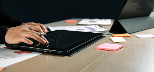 A close-up view of hands working on a digital tablet for data analysis, surrounded by colorful sticky notes and charts on a modern office desk, showcasing productivity.Noogenesis