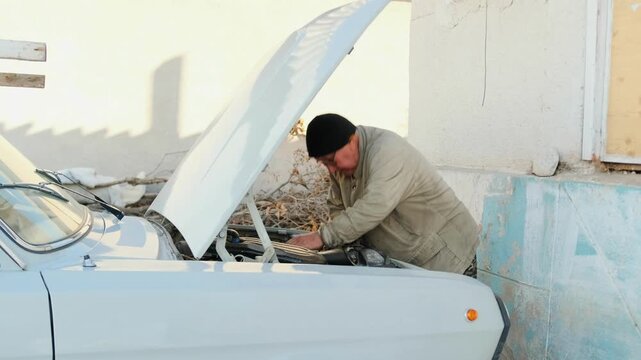 Elderly man repairing vintage car outdoors - senior man working under open hood near sunlit garage - wearing winter coat and black beanie - oily hands checking battery cables - spark plug and