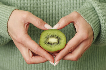Female hands holding kiwi slice in heart shape. Green sweater background. Healthy fruit concept. Fresh organic kiwi closeup. Natural vitamins and diet food. Love healthy lifestyle concept.