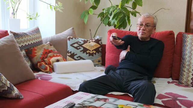 Senior Citizen Unwinds On Comfy Sofa With Newspaper - Older Individual Relaxes With Remote Control And Newspaper - Retired Person Sprawled On Vibrant Sofa Savoring Television And Calming Atmosphere