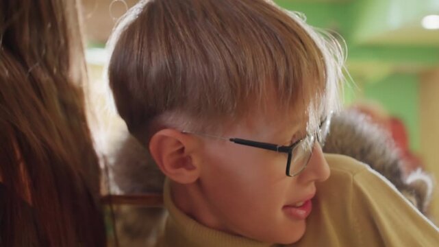 young caucasian boy reading at table wearing glasses and cozy sweater, leaning beside adult companion warm green interior lighting, soft closeup shots show focused expression, whispering and glancing