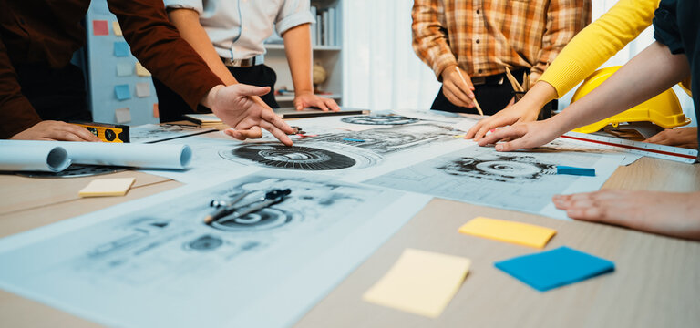 A diverse team engages in collaborative brainstorming while reviewing detailed blueprints and graphics on a table, highlighting innovation and teamwork in a modern office environment. SACTR - Powered by Adobe