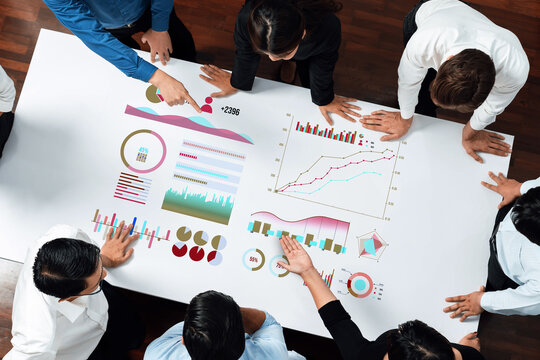 A diverse team of professionals engages in a lively discussion as they analyze various charts and graphs on a large table. The atmosphere is collaborative and focused. Axiom - Powered by Adobe