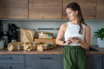 Young woman checks grocery delivery using a mobile app while standing in the kitchen. Concept of app-based meal kit subscription and digital grocery planning for home cooking.