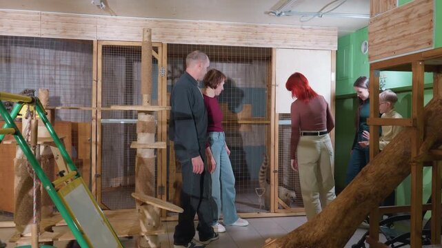 Volunteers inspect wooden animal enclosure inside small team measures frames, uses ladder and tools, cats and dog wander, timber structure and DIY carpentry, concentrated collaborative mood.