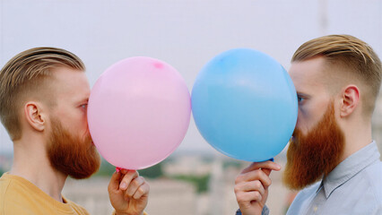 Two identical bearded men face each other while holding pink and blue balloons in front of their faces creating a humorous mirrored effect with a soft urban background and pastel aesthetic