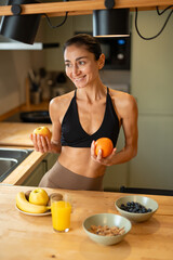 Athletic woman holding fresh apple and orange while preparing healthy breakfast in modern kitchen,...