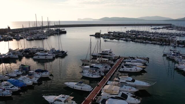 Enjoy stunning aerial views of the marina in Alghero, Sardinia, featuring boats and yachts against a backdrop of historic architecture and the beautiful coastline.