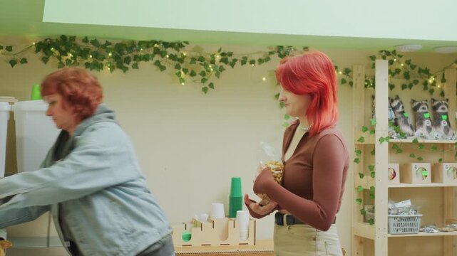 white women playful banana handoff in shop older volunteer quickly passes bananas to young customer at refill counter, jars and bulk bins on shelves, upbeat banter, accidental fumble avoided, bright