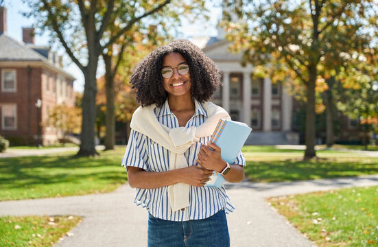 Happy pretty female African American student walking outside university. Smiling cheerful girl model with curly hair wearing glasses, holding notebooks looking at camera outdoors, portrait.