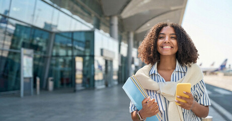 Happy foreign college student holding smartphone standing in airport. Smiling African American girl...