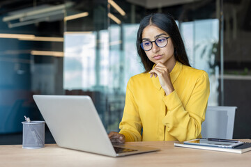 Serious young Indian businesswoman working in the office, sitting at the desk, looking thoughtfully and concentratedly at the laptop screen