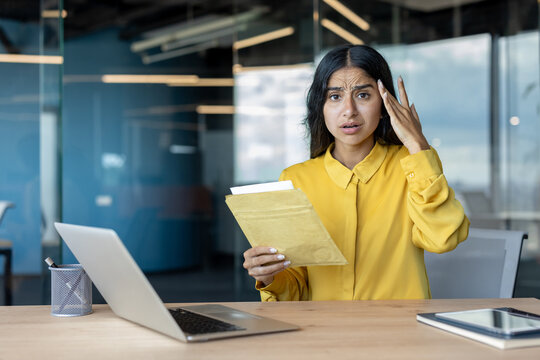 Portrait of a worried young Indian woman sitting at a desk in the office, holding a letter in an envelope and holding her head in shock