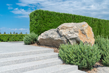 Large boulder in city park. Big stone near stone granite staircase in landscape design. Fragment of beautiful garden against backdrop of trimmed natural border and blue sky. © IhorStore