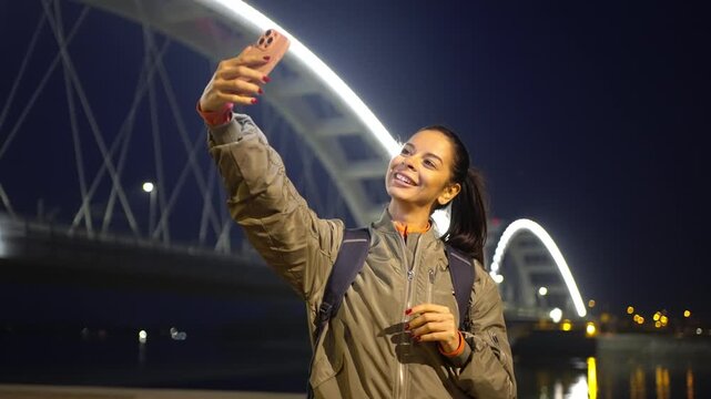 Young woman taking a selfie at night with a bridge illuminated in the background