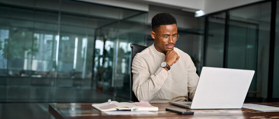 Young male businessman looking at computer thinking on online investment or software technology. Busy professional African business man company employee using laptop working at office.