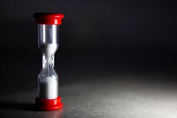 Minimal still life of a small hourglass with red caps, white sand flowing on a dark background, symbolizing time, deadline, countdown, urgency, productivity and focus.
