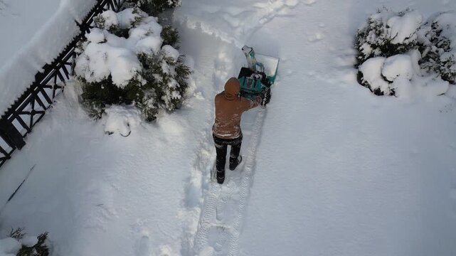 Strong man pushes modern battery-powered snow blower along narrow path covered with white snow. Concentrated male in padded jacket and warm gloves removes snow from walkway in city