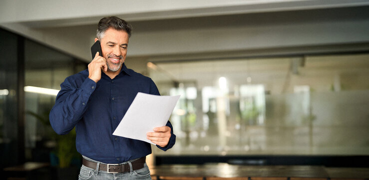 Happy busy mature older business man holding papers making call at work. Smiling middle aged senior professional businessman executive looking at documents talking on cell phone standing in office.