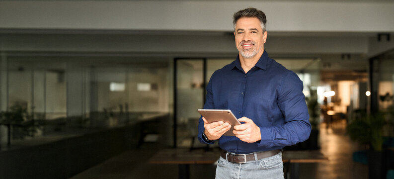 Portrait of happy 50 years old business man executive standing in office using digital tablet. Middle aged businessman manager business owner working on professional finances project looking at camera