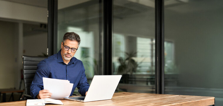 Senior mature male account bank manager executive working on laptop checking paper financial report in office. Busy middle aged professional business man using laptop computer reading document at work