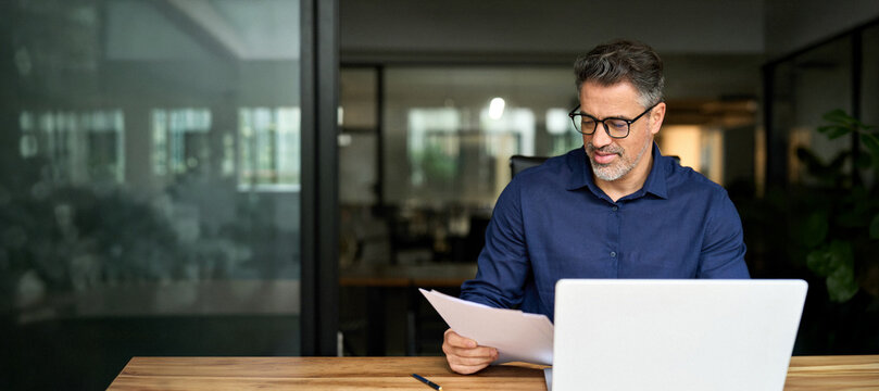 50 years old man executive manager or business owner reading paper working on computer. Busy older professional businessman entrepreneur using laptop holding bill corporate document in office.