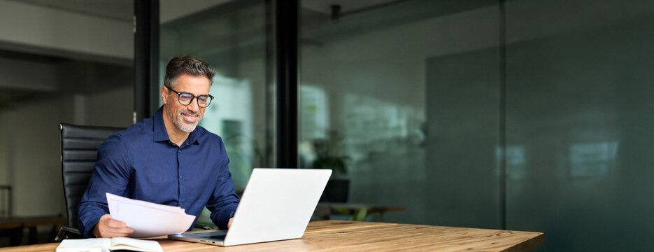 Smiling man executive manager or business owner of middle age reading paper working on computer. Happy busy old professional businessman entrepreneur using laptop holding corporate document in office.
