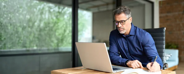 Fototapeten Farbiges Glas 50 years old business man company manager investor working on computer technology sitting at office desk. Busy mature middle aged professional businessman executive using laptop at work writing notes.  © insta_photos
