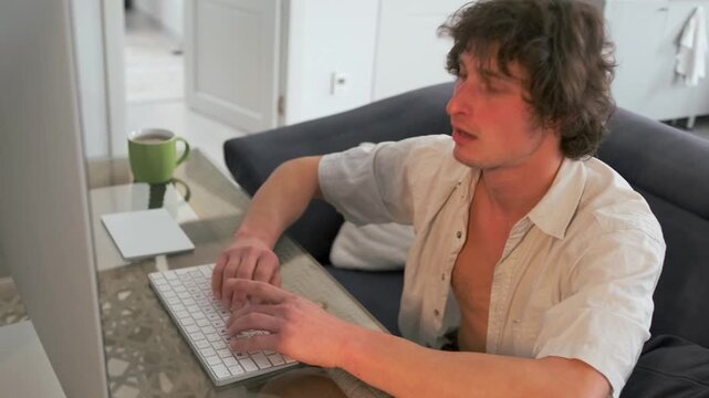 Caucasian man at compact studio desk using mouse and keyboard, engaged in creative editing session with large monitor, mug and notepad nearby, warm indoor lighting, professional focus and