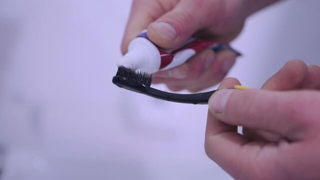 Toothbrush receiving white paste carefully, Gentle squeezing of small toothpaste tube for brushing, Detailed view of hands carefully dispensing toothpaste onto toothbrush in bathroom environment
