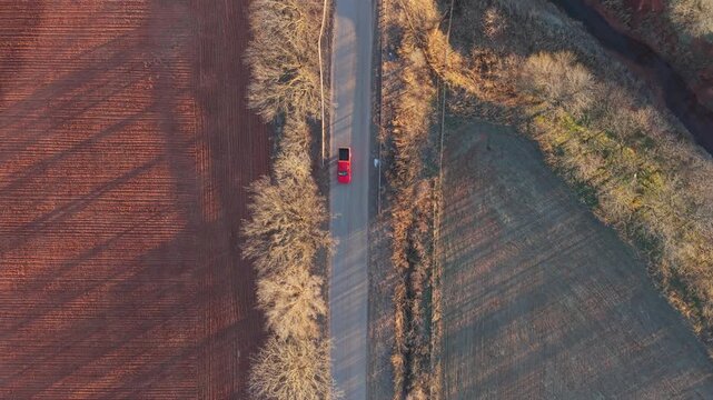 Top-down aerial view of a red pickup truck driving along a rural Oklahoma road between red dirt farmland and winter fields. Golden hour light, bare trees, and quiet countryside textures.
